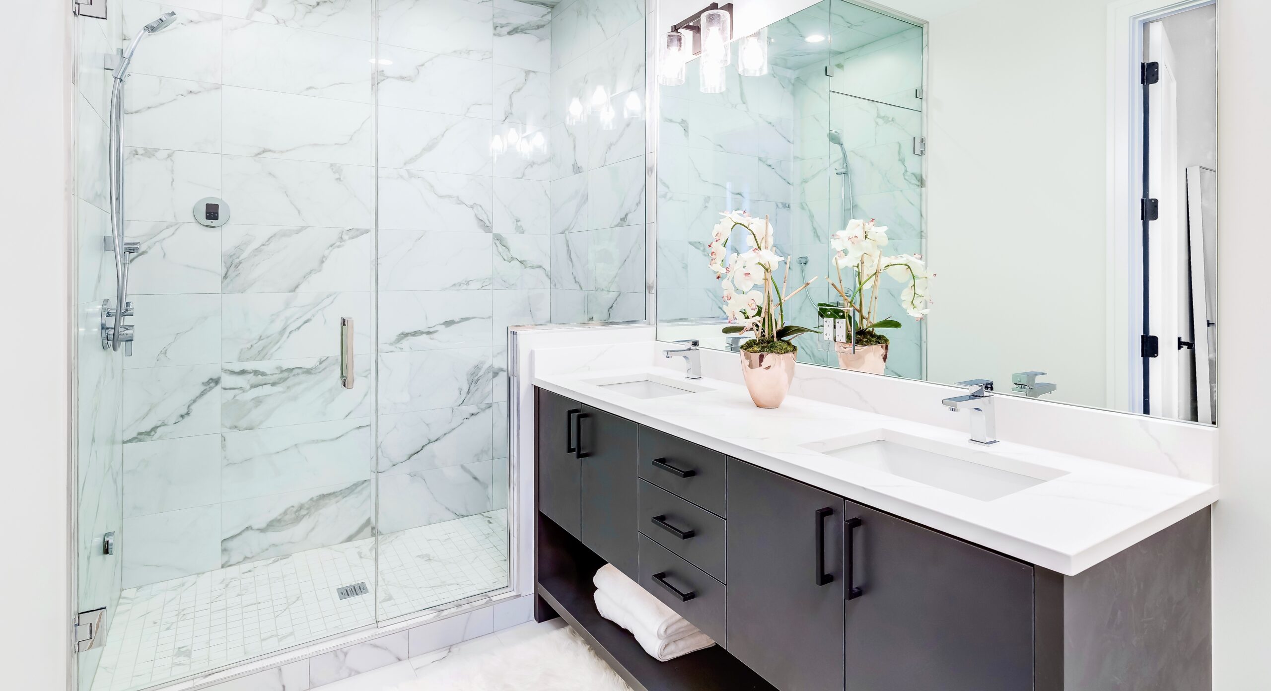 CHICAGO, IL, USA - FEBRUARY 8, 2020: A beautiful bathroom with a dark vanity and white granite counter top. A plant and towels sit on the vanity. The shower is lined with marble tiles. Lights on.